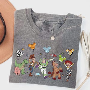 Może przedstawiać: Szara koszulka Comfort Colors z okrągłym dekoltem i kolorową grafiką postaci z Toy Story. Projekt zawiera Chudego, Buzz'a Astrala, Jessie i inne postacie z balonami w kształcie Myszki Miki. Widoczny jest r&oacute;wnież srebrny naszyjnik z wisiorkiem.