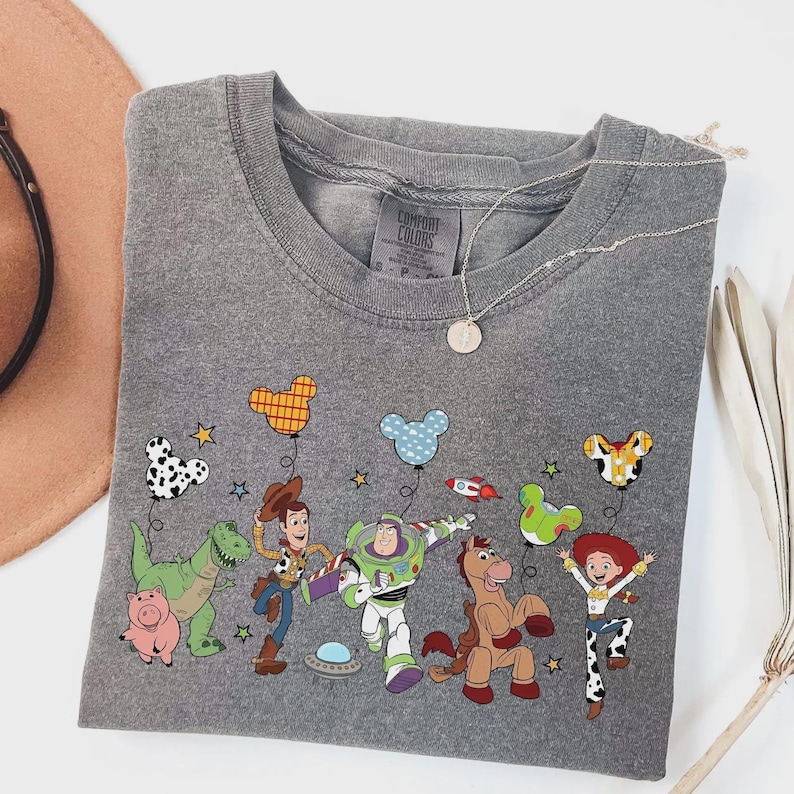 Może przedstawiać: Szara koszulka Comfort Colors z okrągłym dekoltem i kolorową grafiką postaci z Toy Story. Projekt zawiera Chudego, Buzz'a Astrala, Jessie i inne postacie z balonami w kształcie Myszki Miki. Widoczny jest r&oacute;wnież srebrny naszyjnik z wisiorkiem.