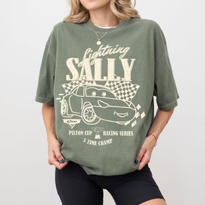 May include: Olive green t-shirt with the text "Lightning Sally" above a cartoon car graphic. The graphic includes a checkered flag and the text "Piston Cup Racing Series 5 Time Champ".
