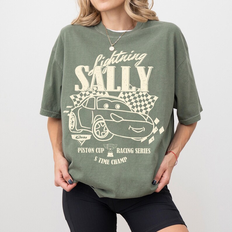 May include: Olive green t-shirt with the text "Lightning Sally" above a cartoon car graphic. The graphic includes a checkered flag and the text "Piston Cup Racing Series 5 Time Champ".