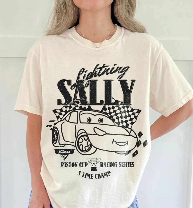 May include: Cream-colored t-shirt featuring a black graphic of a car with the text "Lightning Sally" above it. The design includes checkered flags and the words "Piston Cup Racing Series" and "5 Time Champ".