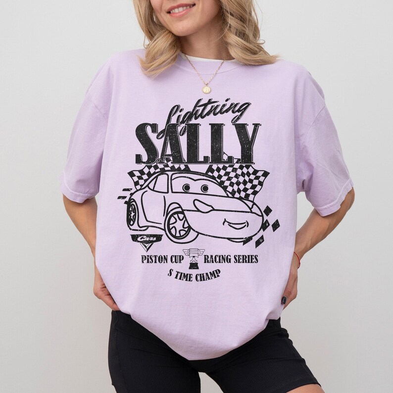 May include: A light purple t-shirt featuring a black and white graphic of the character Sally from the movie Cars. The graphic includes the text "Lightning Sally", a race car, and the words "Piston Cup Racing Series 5 Time Champ".