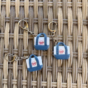 May include: Three miniature Trader Joe's shopping bag keychains. Each keychain features a white bag with blue accents and the Trader Joe's logo in red. The keychains are attached to gold-colored metal rings and chains.