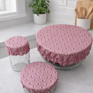 May include: Pink fabric bowl covers with a bee pattern. The covers are in three sizes, designed to fit various bowls and jars. The fabric has a ruffled edge. The covers are on a white countertop.