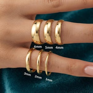 Puede incluir: Seis anillos de tono dorado con superficies lisas y pulidas, mostrados en los dedos. Los anillos están etiquetados con anchos de 6mm, 5mm, 4mm, 3mm, 2mm y 1mm, indicando sus respectivos grosores en milímetros para una comparación clara.