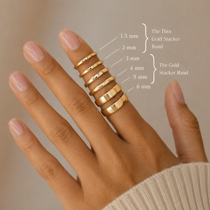May include: A series of six smooth gold bands stacked on a finger, labelled with widths of 1.5 mm, 2 mm, 3 mm, 4 mm, 5 mm, and 6 mm. The text identifies the thinner rings as The Thin Gold Stacker Band and the wider rings as The Gold Stacker Band.