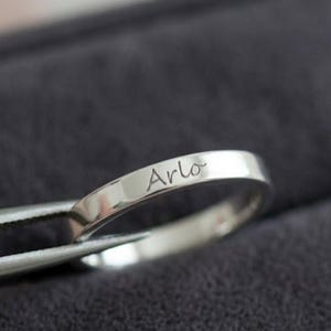 Puede incluir: Un anillo de metal plateado pulido con el nombre Arlo grabado en letra cursiva en la superficie exterior. El anillo está sujeto por unas pinzas de metal, mostrando el acabado liso y reflectante de la banda y las letras claramente visibles.