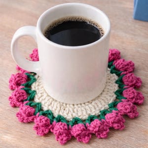 May include: A white ceramic mug filled with black coffee sits on a handmade crochet coaster. The coaster features a cream-colored center with a border of pink flower-like shapes and green stems. The mug has a curved handle and is placed on a wooden surface.