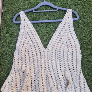 Puede incluir: Un top de ganchillo beige sin mangas con un escote en V profundo y un dobladillo con volante acampanado. La prenda est&aacute; confeccionada con un patr&oacute;n de punto calado en toda su superficie, creando un aspecto texturizado y ligero. Se muestra en una percha gris.