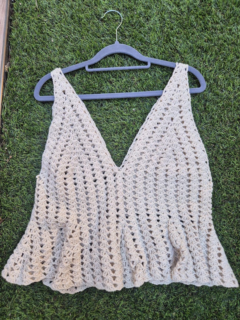 Puede incluir: Un top de ganchillo beige sin mangas con un escote en V profundo y un dobladillo con volante acampanado. La prenda est&aacute; confeccionada con un patr&oacute;n de punto calado en toda su superficie, creando un aspecto texturizado y ligero. Se muestra en una percha gris.