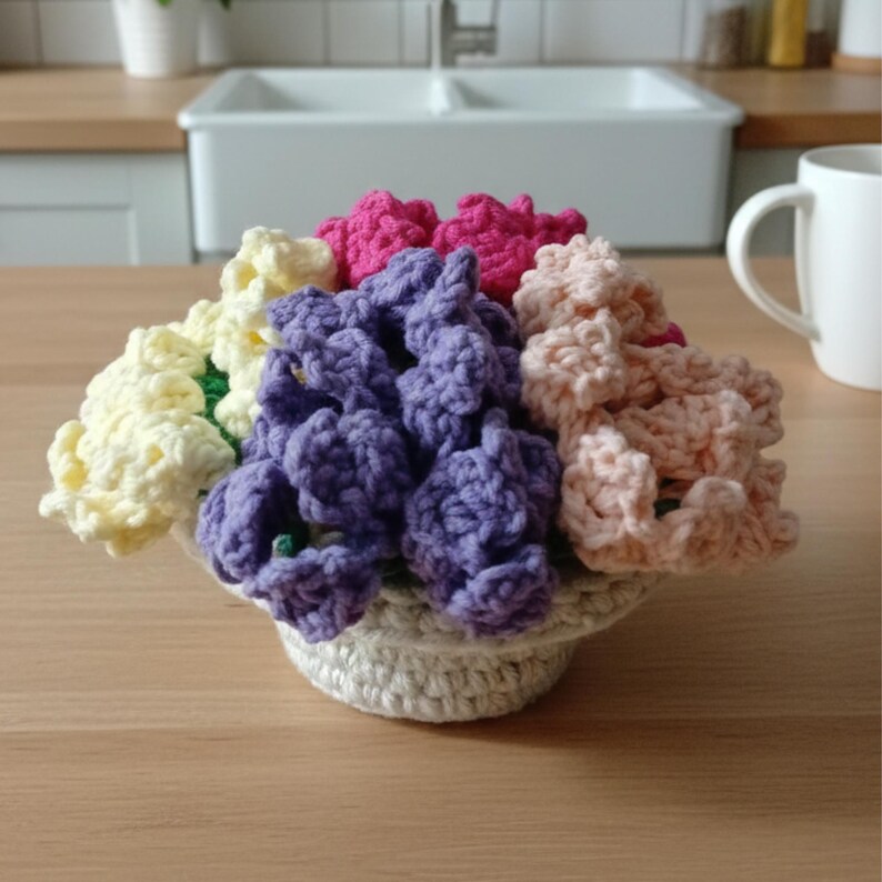 May include: A crocheted flower arrangement in a small, off-white basket. The flowers are in shades of yellow, pink, purple, and peach. The arrangement is sitting on a wooden surface, with a white mug and kitchen sink in the background.