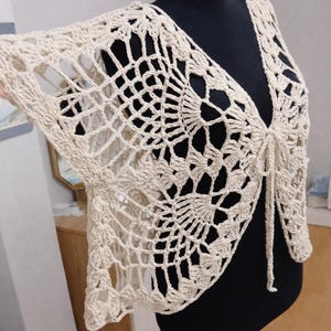 May include: A cream-colored, open-weave crochet shrug displayed on a black mannequin. The shrug features a V-neck and tie closure, with a delicate, lacy pattern. The sleeves extend to the elbows, showcasing the intricate design. The garment is a handmade, bohemian style.