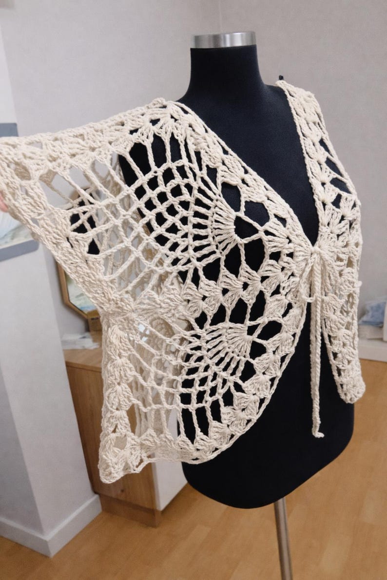 May include: A cream-colored, open-weave crochet shrug displayed on a black mannequin. The shrug features a V-neck and tie closure, with a delicate, lacy pattern. The sleeves extend to the elbows, showcasing the intricate design. The garment is a handmade, bohemian style.