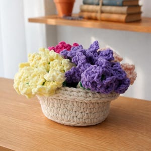 May include: A crocheted flower arrangement in a cream-colored basket. The flowers are in shades of yellow, pink, purple, and peach. The basket is round and has a textured, woven appearance. The arrangement sits on a wooden surface.