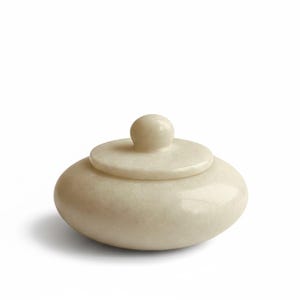 May include: An off-white, oval jar with a lid. The lid has a small, round knob. The jar and lid have a smooth, polished finish. The jar is likely made of stone or ceramic. The jar's dimensions are approximately 10 cm in diameter.