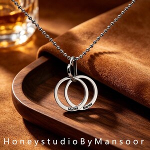 May include: A silver-toned necklace featuring a delicate ball chain and a pendant composed of two interlocking circular rings. Each ring is engraved with the name Marley in a clean, black sans-serif font. The pendant hangs from a small heart-shaped loop.