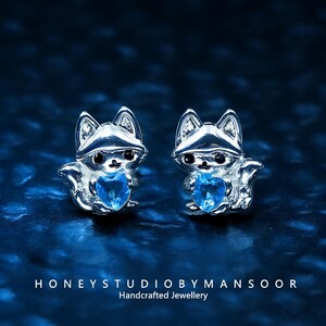 Puede incluir: Pendientes de botón plateados con forma de zorro, cada uno con una gema azul en forma de corazón. Los pendientes se presentan sobre un fondo azul oscuro. El texto "HONEYSTUDIOBYMANSOOR Handcrafted Jewellery" está en la parte inferior.