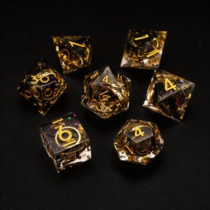 Puede incluir: Un conjunto de siete dados poliédricos en negro y dorado. Los dados están hechos de resina transparente con números dorados y elementos decorativos internos. Los dados están dispuestos sobre una superficie negra.