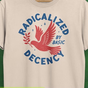 Könnte beinhalten: Elfenbeinfarbenes T-Shirt mit einer Grafik einer roten Taube, die einen Olivenzweig trägt, umgeben von den blauen Worten "RADICALIZED BY BASIC DECENCY". Das Shirt hängt an einem Holzkleiderbügel.