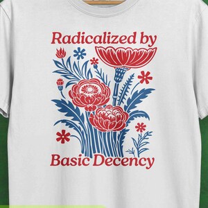 Può includere: T-shirt bianca con un design floreale rosso e blu e la scritta "Radicalized by Basic Decency". La parola "WHITE" è stampata in basso. La maglietta è appesa a una gruccia di legno.