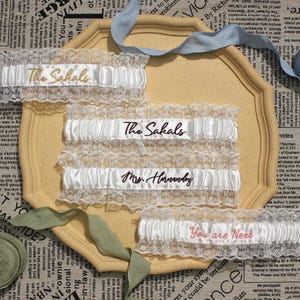 May include: Four white satin wedding garters with lace trim, each with embroidered text. The text includes "The Sahals", "Mrs. Hornsby", and "You are Next". The garters are displayed on a decorative, light yellow tray.
