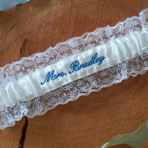 May include: Ivory satin wedding garter with delicate white lace trim. The garter features the embroidered name "Mrs. Bradley" in blue script. A light blue ribbon and a green ribbon are visible in the background.