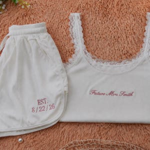 Könnte beinhalten: Weißes Pyjama-Set mit Tanktop und Shorts. Das Tanktop hat eine Spitzenborte und den gestickten Text "Future Mrs. Smith". Die Shorts haben den gestickten Text "EST. 8/22/26". Das Set wird auf einer pfirsichfarbenen Oberfläche präsentiert.