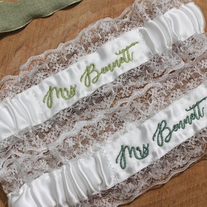May include: Two white satin and lace garters with the embroidered name "Mrs. Bennett" in green script. A green ribbon is visible in the upper left corner. The garters are on a wooden surface.
