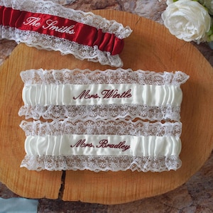 Può includere: Una collezione di giarrettiere da sposa. Una giarrettiera rossa con "The Smiths" ricamato in bianco e due giarrettiere bianche con finiture in pizzo, ciascuna ricamata con un nome in bordeaux. Le giarrettiere sono esposte su una superficie di legno.