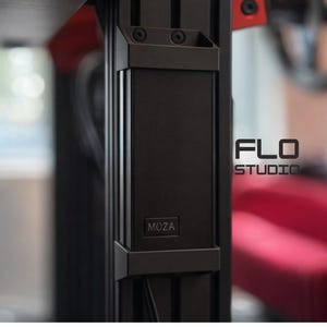 May include: A close-up of a black metal structure with the text "MOZA" on the front. The structure is part of a larger system, with the text "FLO STUDIO" visible in the background. A red sofa is also visible.