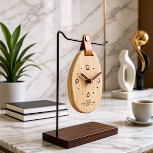 Puede incluir: Un reloj ovalado de madera con una correa de cuero marrón, manecillas negras y una base marrón oscuro. El reloj tiene los números 12, 3, 6 y 9 marcados con puntos. El reloj está suspendido de un soporte de metal negro.