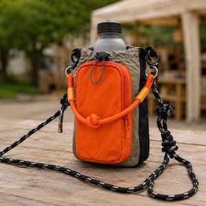May include: A water bottle holder bag featuring a sage green main compartment with a bright orange zippered front pocket. The bag includes a black drawstring closure, a thick orange rope handle, and a long black and white patterned rope shoulder strap.