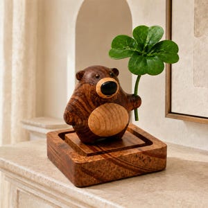 Puede incluir: Una figura de oso de madera tallada con cuerpo marrón oscuro, vientre circular de color beige claro y nariz negra. El oso está sobre una base cuadrada de madera y sostiene un tallo de trébol verde de cuatro hojas en su pata derecha.
