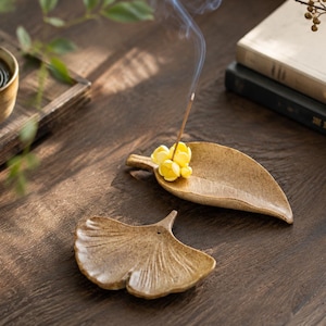May include: Two ceramic incense holders with a speckled tan finish. One holder is shaped like an elongated leaf and holds a burning incense stick with a small yellow flower accent. The other holder is shaped like a ginkgo leaf with visible veins.