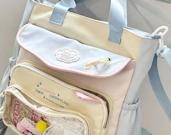 May include: A pastel-colored tote bag with a light blue handle and shoulder strap. The bag features a front pocket with a clear window displaying decorative items. Text on the bag reads "UMIA THINKING ONDAYLUC + EXCLUSIVE 2 TO TIME".