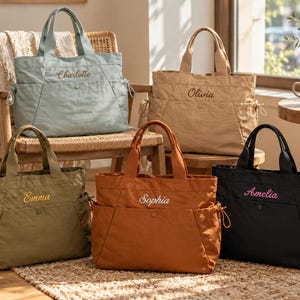 May include: Five tote bags in sage green, tan, olive green, rust orange, and black. Each bag features two top handles, side pockets with drawstrings, a center snap button, and embroidered names: Charlotte, Olivia, Emma, Sophia, and Amelia.