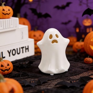Puede incluir: Una figura de fantasma de cerámica blanca con una expresión de sorpresa, rodeada de calabazas naranjas y decoraciones de murciélagos negros. La palabra "YOUTH" es visible en una pila de libros blancos. Decoración temática de Halloween.