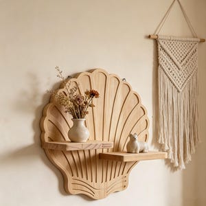 May include: A wooden seashell-shaped wall shelf with two shelves. The shelf holds a small vase with dried flowers and a ceramic figurine. A macrame wall hanging is to the right.
