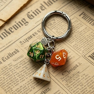 Op de afbeelding: Een zilveren sleutelhanger met drie kleurrijke dobbelsteenbedels. De bedels omvatten een groene D20, een oranje D6 en een witte D4. De dobbelstenen hebben goudkleurige cijfers. De sleutelhanger heeft een kleine hartbedel met "made with love".