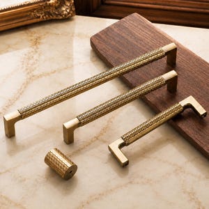 May include: Three brass-toned metal cabinet pulls of varying lengths and one matching cylindrical knob. Each piece features a textured, repeating scale-like pattern along the center and smooth, angular mounting brackets at the ends.