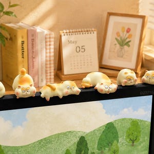 Könnte beinhalten: Ein Set aus sechs kleinen, cartoonartigen Katzenfiguren in verschiedenen Posen, gefärbt in Creme- und Orangetönen. Diese dekorativen Ornamente sitzen auf der Oberkante eines schwarzen Computermonitors und zeigen jeweils einen eigenen Gesichtsausdruck.