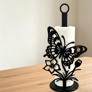 May include: Black metal paper towel holder featuring a butterfly and floral design. The butterfly has intricate cut-out details. The holder has a circular base and a handle at the top. A roll of white paper towels is in the center.