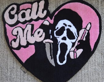 Puede incluir: Una alfombra de mechones en forma de corazón con fondo rosa y el texto negro Call Me. El diseño incluye una figura blanca similar a un fantasma con capucha negra que sostiene un cuchillo plateado y un auricular de teléfono rosa sobre un corazón.