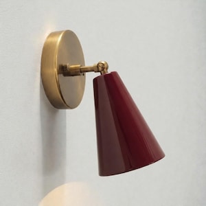 Puede incluir: Una lámpara de pared con acabado de latón y una pantalla cónica de color rojo oscuro. La lámpara tiene una base circular y un brazo ajustable. La pantalla está inclinada hacia abajo y la lámpara está montada en una pared blanca.