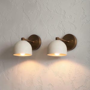 Könnte beinhalten: Zwei Wandleuchten mit weißem, kuppelförmigem Schirm und warmgoldenem Innenraum. Die Leuchten sind an einer kreisförmigen, bronzefarbenen Basis mit passendem Arm befestigt. Die Lichter sind eingeschaltet und erzeugen einen warmen Schein.