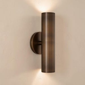 Puede incluir: Aplique de pared cilíndrico de color bronce con una placa trasera circular. El aplique emite luz desde la parte superior e inferior, creando un brillo cálido. La luminaria está montada en una pared de color claro.