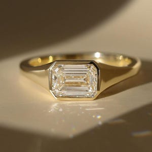 May include: A gold ring featuring a single emerald-cut clear gemstone set in a smooth bezel setting. The band is polished yellow gold with a tapered design that leads to the central rectangular stone, creating a clean and reflective appearance.