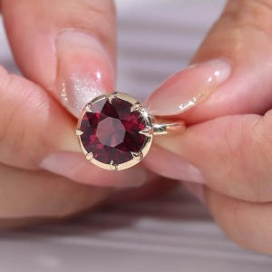Pode incluir: Um anel com uma grande pedra preciosa redonda de cor vermelho-profundo, montada numa banda de ouro polido. A pedra é fixada por várias garras que se estendem sobre a borda da gema, criando um engaste seguro e brilhante no metal.