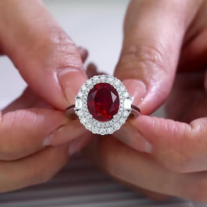 May include: A silver-toned ring featuring a large, oval-cut deep red gemstone at the centre. The stone is surrounded by a double halo of small, round, clear white diamonds, with two baguette-cut diamonds set on the sides of the band.