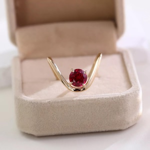 May include: A V-shaped gold ring featuring a single round-cut deep red gemstone set in a four-prong setting at the centre of the band. The polished gold band curves upwards into a chevron shape, highlighting the vibrant red stone at the base.